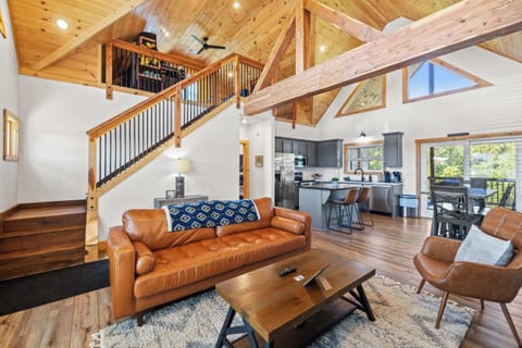 Spacious living room with plush seating, soaring wood accents, and stairs leading to the game loft. Sunlight pours through the tall windows, filling the open dining and kitchen areas with warmth—perfect for gathering, relaxing, and making memories.