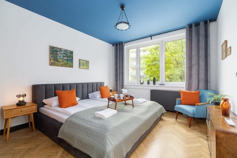 A cozy bedroom with a large bed covered in gray bedding and orange accent pillows. There is a bright window with curtains and a bedside table with a lamp. The wooden floor adds warmth to the room.