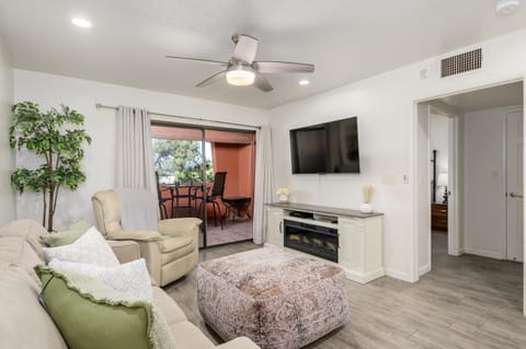 Living room with beige sofa, leather recliner, patterned ottoman, wall-mounted TV, and sliding glass door to balcony.