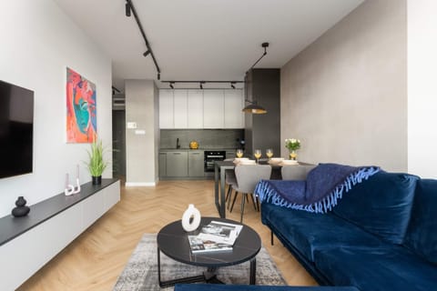 A modern, open-plan living room with a blue sofa, a dining table with chairs, and a large window letting in natural light.