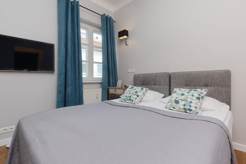 The studio features a cozy bed with soft gray bedding and decorative patterned pillows, placed near a window with long blue curtains.
