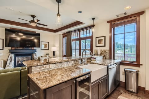 A modern kitchen and living area with granite countertops, farmhouse sink, large windows, wall-mounted TV, ceiling fan, and wooden trim. The layout includes a counter seating area and adjacent seating room.