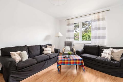 A cozy and bright living room with two black sofas, a colorful coffee table cover, and a large window filling the space with natural light.


