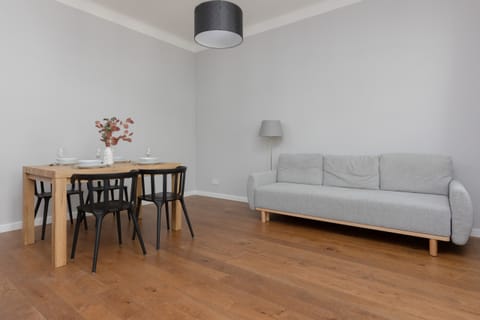 A bright and spacious living area with a comfortable gray sofa, wooden flooring, and a dining table with chairs, perfect for relaxing or entertaining.
