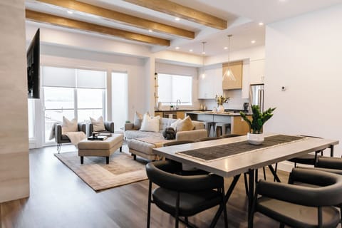 This modern open-concept space features a cozy beige sectional, sleek dining area, and a stylish kitchen with stainless steel appliances. Exposed wooden beams and natural light create a warm, inviting atmosphere perfect for relaxation.