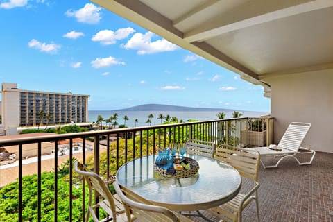 Furnished lanai featuring a glass-top dining table, cushioned seating, and sweeping ocean views framed by lush tropical greenery.