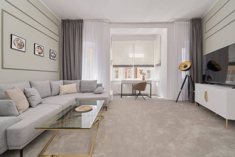 A spacious living room with a gray sofa, a glass coffee table, and beige carpeting, featuring soft natural light from a large window.