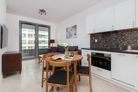 An open-plan kitchen and dining area with white cabinetry, black backsplash tiles, and a wooden dining table for four.

