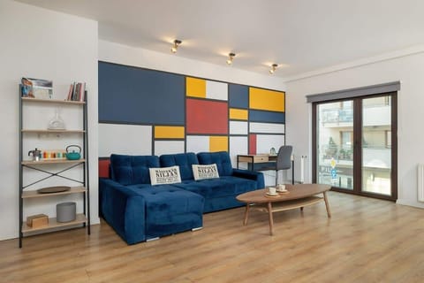 A vibrant living room with a blue sofa, colorful geometric wall decor, a wooden coffee table, and large windows providing natural light.

