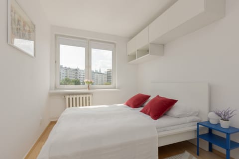 A bright bedroom featuring a double bed with white linens and vibrant red pillows, complemented by a large window with natural light.

