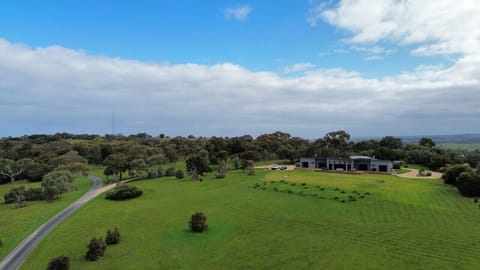 An aerial view of a large, green landscape with a modern house situated among trees and a winding road leading up to it, under a partly cloudy sky.