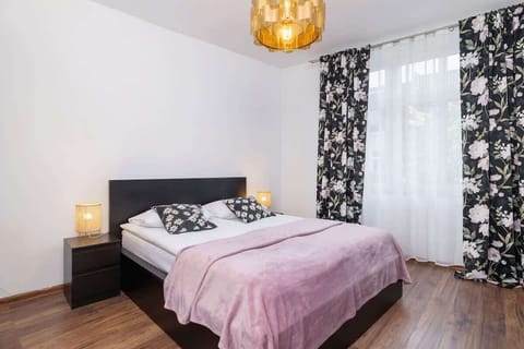 A cozy bedroom with a soft pink duvet, bedside lamp, and floral-patterned curtains creating a relaxing atmosphere.
