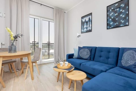 A cozy living room with a blue sectional sofa, a wooden coffee table, and large windows leading to a balcony, filling the space with natural light.

