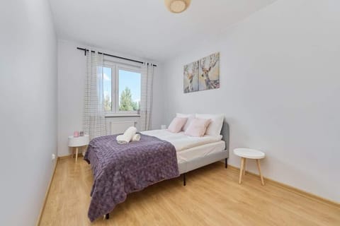A cozy bedroom with a double bed covered in light bedding and a purple throw blanket, featuring a minimalist side table and a large window.

