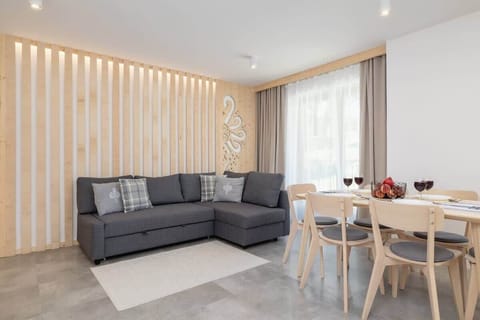A spacious living and dining area with a gray sofa, a dining table, and wooden wall accents, creating a modern yet cozy ambiance.
