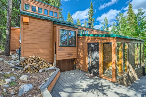 A modern wooden cabin with large windows, set in a forested area. A stack of firewood is visible near the entrance. The scene is sunny with a blue sky and scattered clouds.