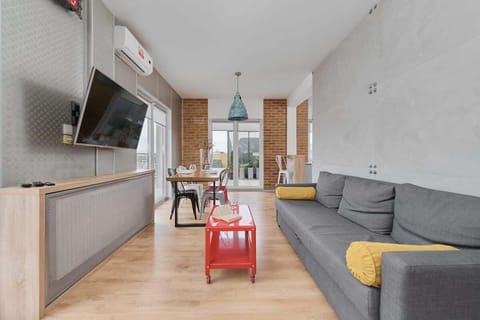 A bright living room with a gray sofa, a red coffee table, and a TV unit, illuminated by natural light from large windows.

