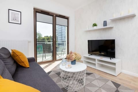 A bright living room with a gray sofa, yellow cushions, wall-mounted TV, and large windows letting in natural light.