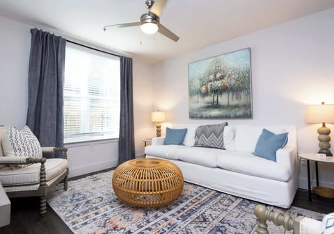 A cozy and bright living room featuring a crisp white sofa, a decorative woven ottoman, and soft blue accent pillows.