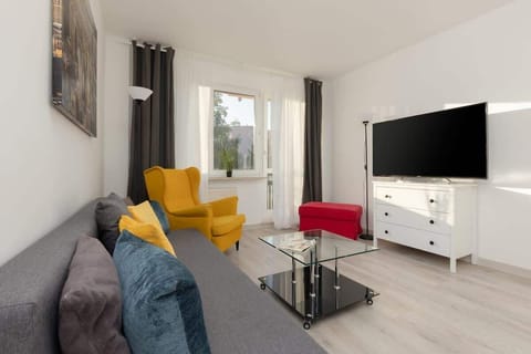 A bright living room with a grey sofa, a yellow armchair, a white sideboard, and a large flat-screen TV. Big windows let in plenty of natural light.
