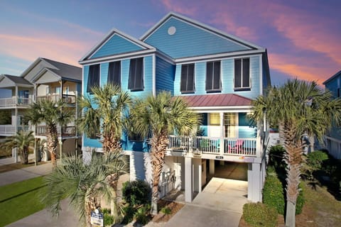 Charming coastal rental with vibrant blue siding, surrounded by swaying palm trees under a stunning sunset sky.