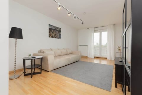 A bright living room featuring wooden floors, a spacious beige sectional sofa, and large windows with plenty of natural light, creating an inviting atmosphere.