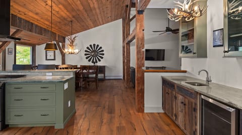 A spacious, rustic kitchen and dining area with wooden ceilings, a green kitchen island, a dining table under a pendant light, and visible living room area. Deer antler light fixtures adorn the space.