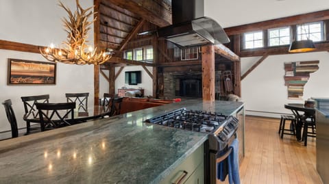 A rustic kitchen and dining area with wooden beams, a stone fireplace, a large antler chandelier, and a kitchen island with a stovetop and oven.