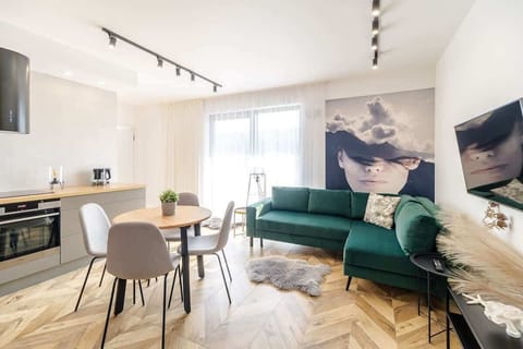 A bright living area with a green sofa, a round dining table, and a large window. A portrait mural adds a stylish accent on the wall.
