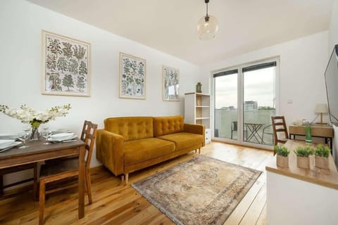 A bright living room with a mustard yellow sofa, a patterned rug, and a dining table in the background.
