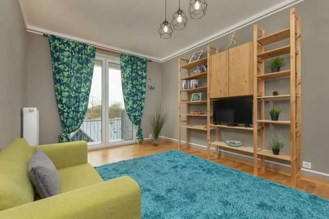 A cozy living room with a green sofa, wooden bookshelves, a blue rug, and large windows with green curtains.
