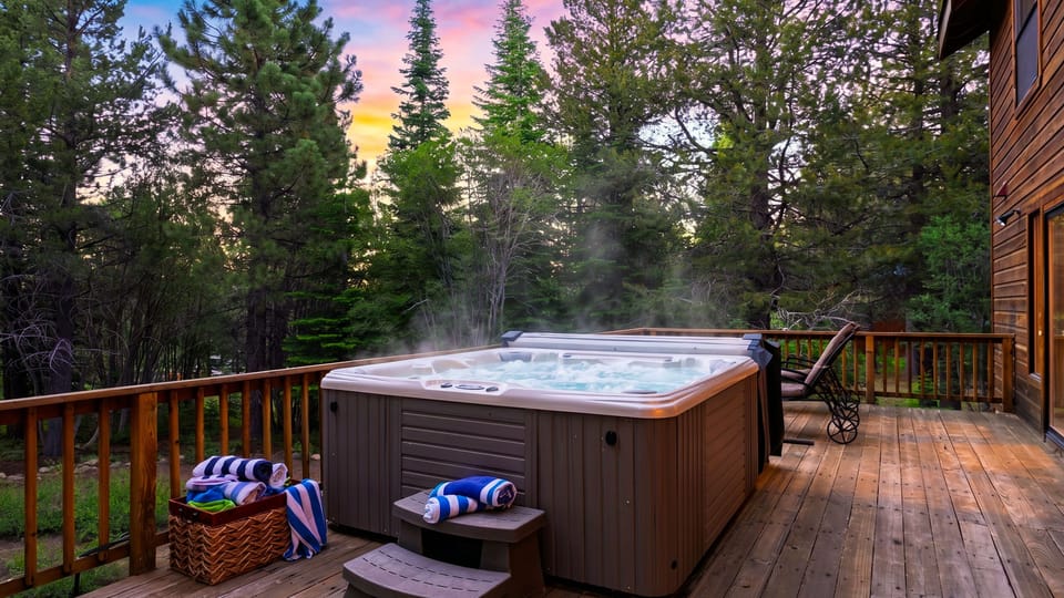 Steaming deck hot tub at sunset, surrounded by tall pines and peaceful mountain scenery.