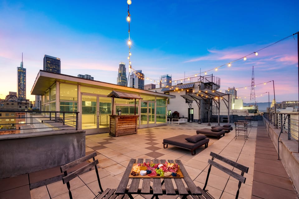 A stylish rooftop patio at dusk, with string lights illuminating a seating area, a bar, and lounge chairs, all set against a stunning city skyline.