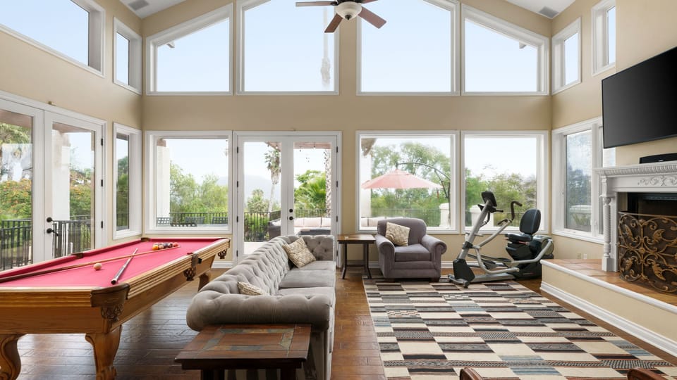 Sun-drenched great room boasting soaring vaulted ceilings, architectural windows, a billiard area, and a fitness corner.