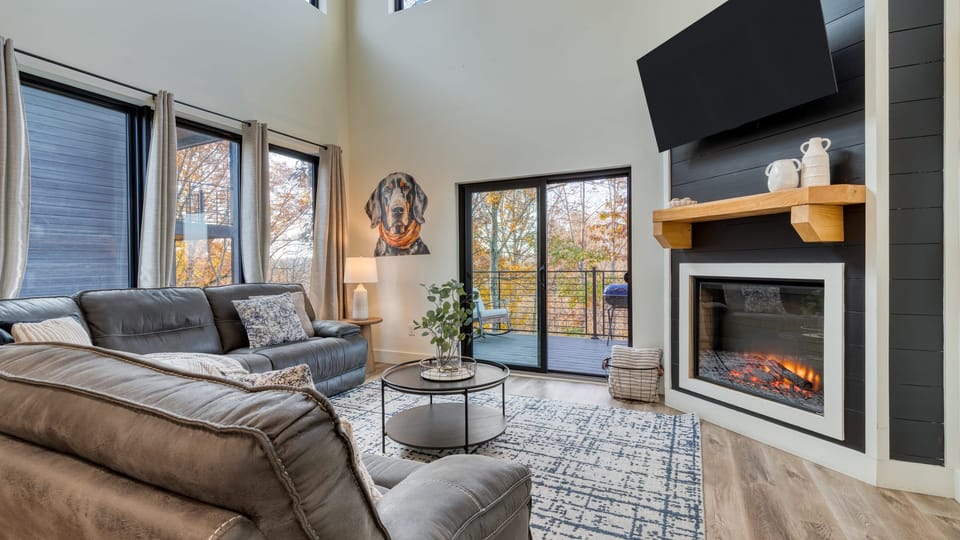 Modern living room featuring a high ceiling, multiple windows, a TV, and a fireplace.