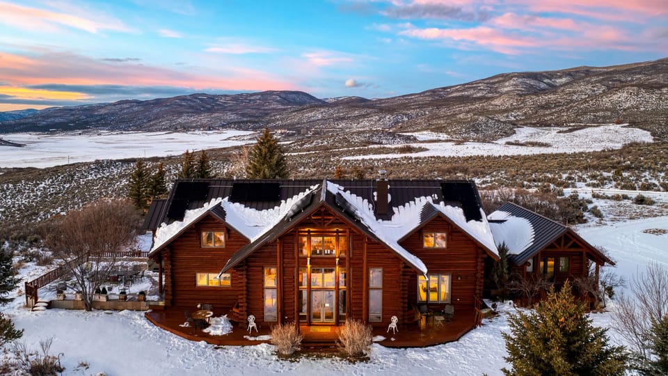 Exterior of a large log cabin home at sunset, covered in snow, with a mountain view.
