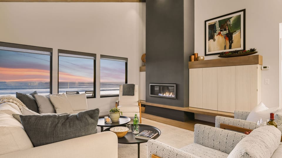 Living room with a cozy seating area featuring a minimalist fireplace and large windows facing the ocean at sunset.