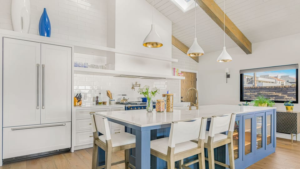 A bright, modern kitchen with white cabinetry, a large blue island, and vaulted ceilings with exposed beams.
