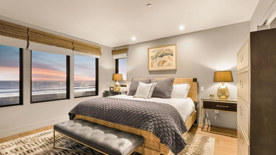 Spacious primary bedroom with a king-sized bed, soft grey tones, and a large window overlooking the water.