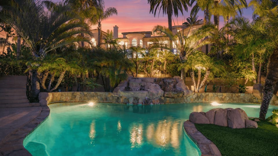 Resort‑style backyard with a glowing pool, lush palms, and a grand estate backdrop.