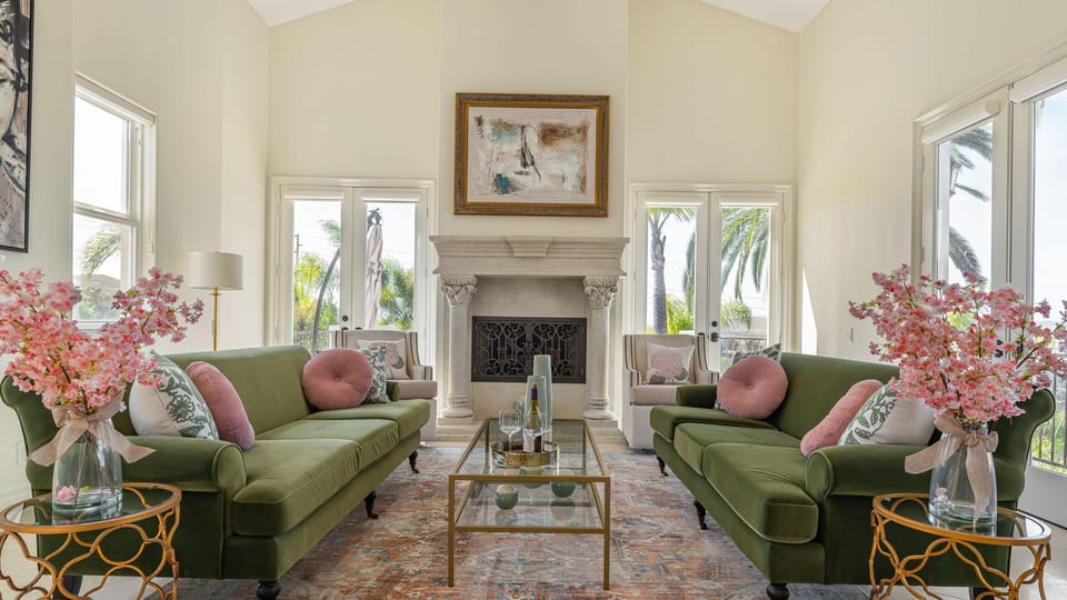 Bright, elegant second living room with green velvet sofas, a stone fireplace, and sun‑filled views framed by tall windows.