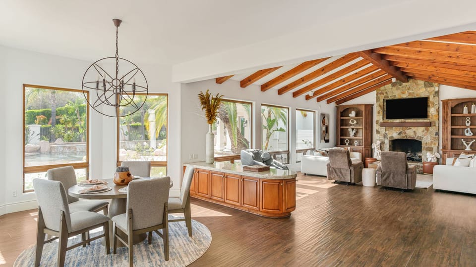 Bright, open living and dining space with vaulted beams, a stone fireplace, and garden views — a warm, airy Temecula retreat.
