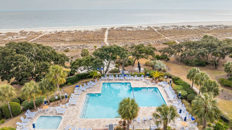 Resort‑style communal pool deck with rows of loungers, palm‑lined paths, and a clear view leading straight to the beach.