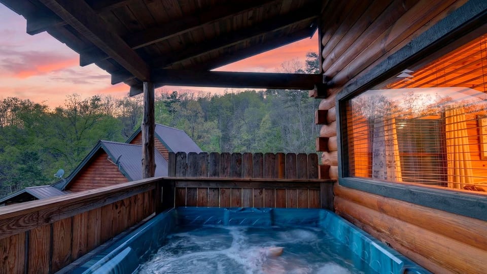 Covered cabin deck with a bubbling hot tub and a colorful sunset over the surrounding forest and nearby cabins.