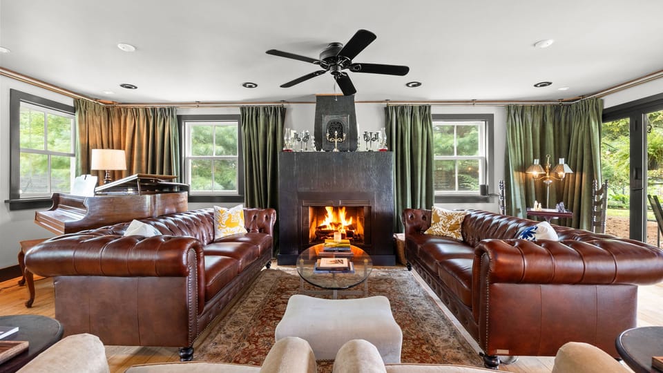 Welcoming living room featuring plush leather sofas, a cozy fireplace, a grand piano, and elegant dark green drapery.