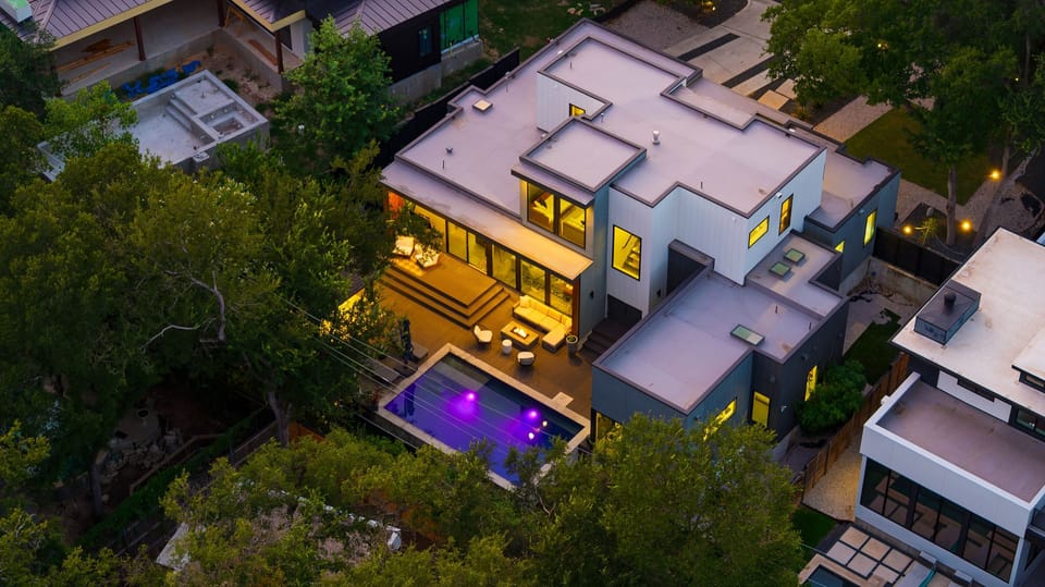 A twilight aerial showcases the modern home, glowing pool, and inviting outdoor lounge tucked among the trees.