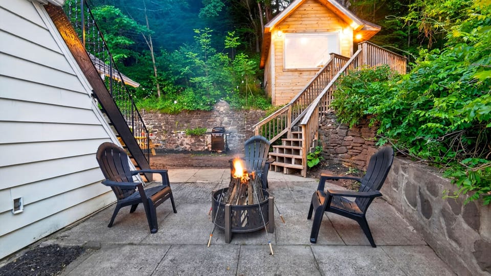 Fire pit patio with Adirondack seating, a nearby grill, and a cozy wooded backdrop.