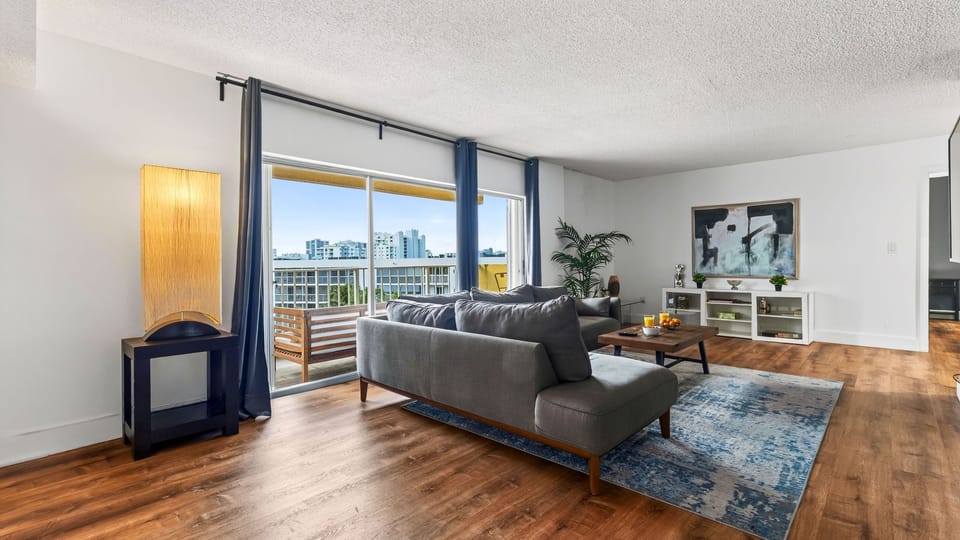A spacious living room opens to a private balcony, with comfortable seating and city views just beyond the sliding doors.