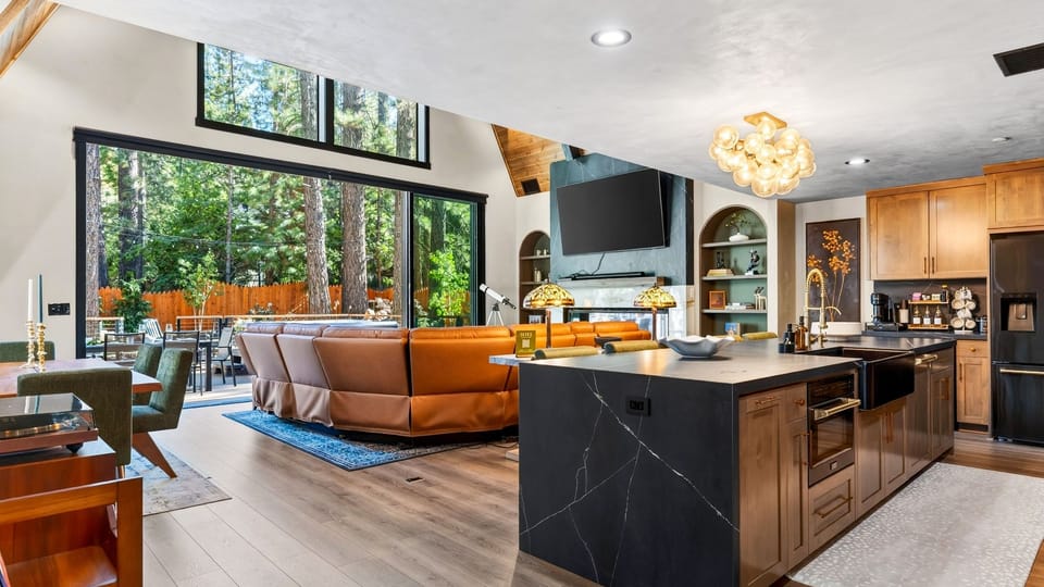 The open-concept great room brings the kitchen and lounge together beneath soaring wood ceilings and expansive forest-facing windows.