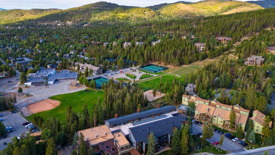 A sweeping aerial view showcases the surrounding mountain town, evergreen forests, and high-country scenery.